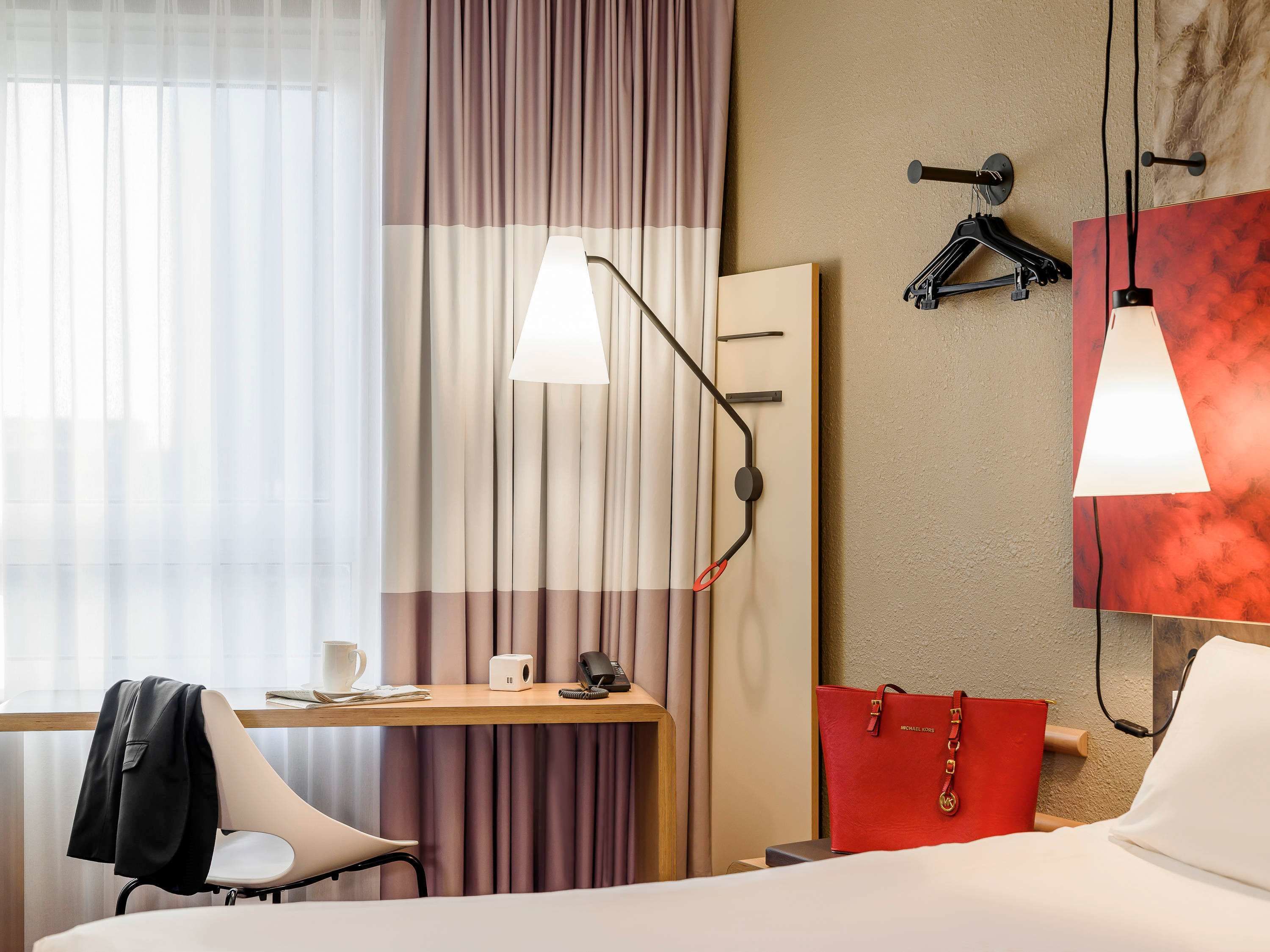 A hotel room with a bed, a desk with a chair, a telephone, a red bag, and two wall-mounted lamps.