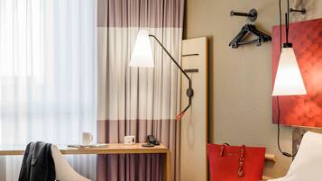 Ein Hotelzimmer mit einem Bett, einem Schreibtisch mit Stuhl, einem Telefon, einer roten Tasche und zwei Wandleuchten.