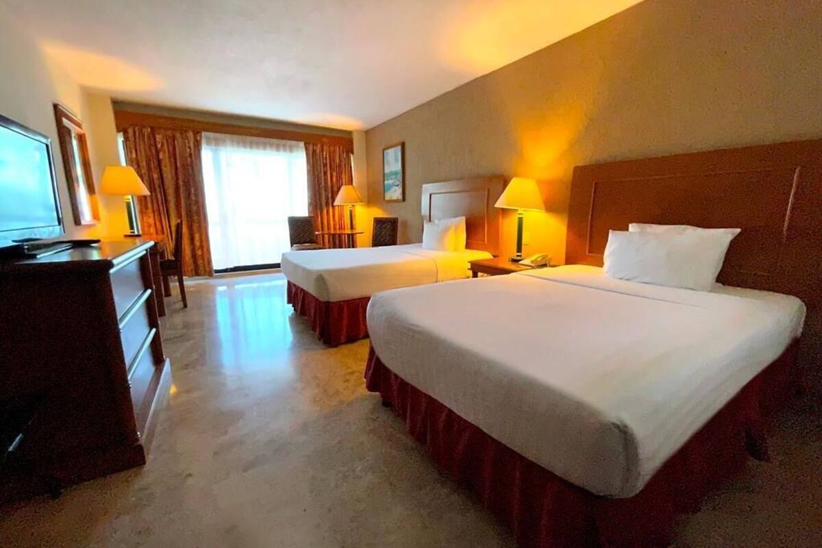 A hotel room with two beds, a desk, a TV, and a window with curtains.