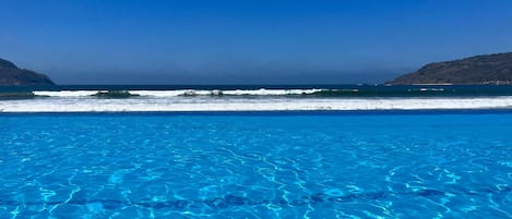 Una piscina azul cristalina con vistas al océano y colinas al fondo.
