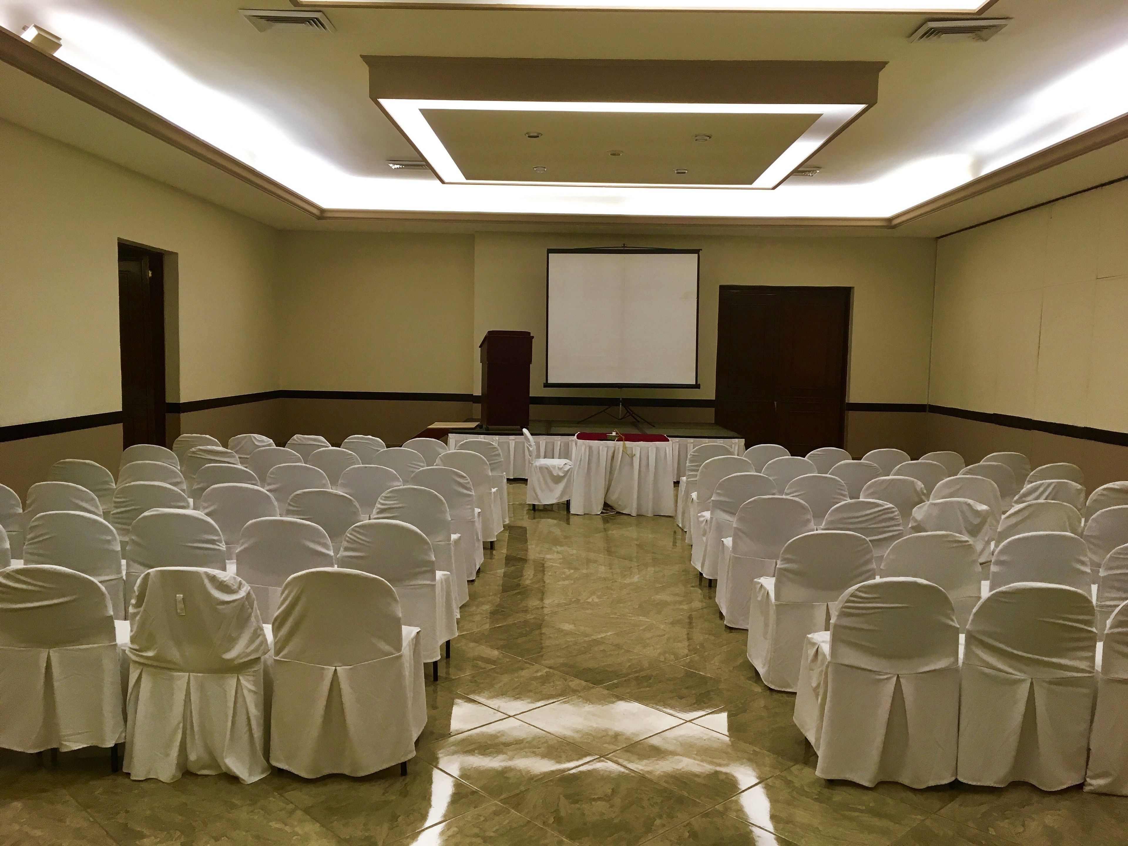 A conference room with rows of chairs covered in white, a podium, a screen, and a tiled floor.