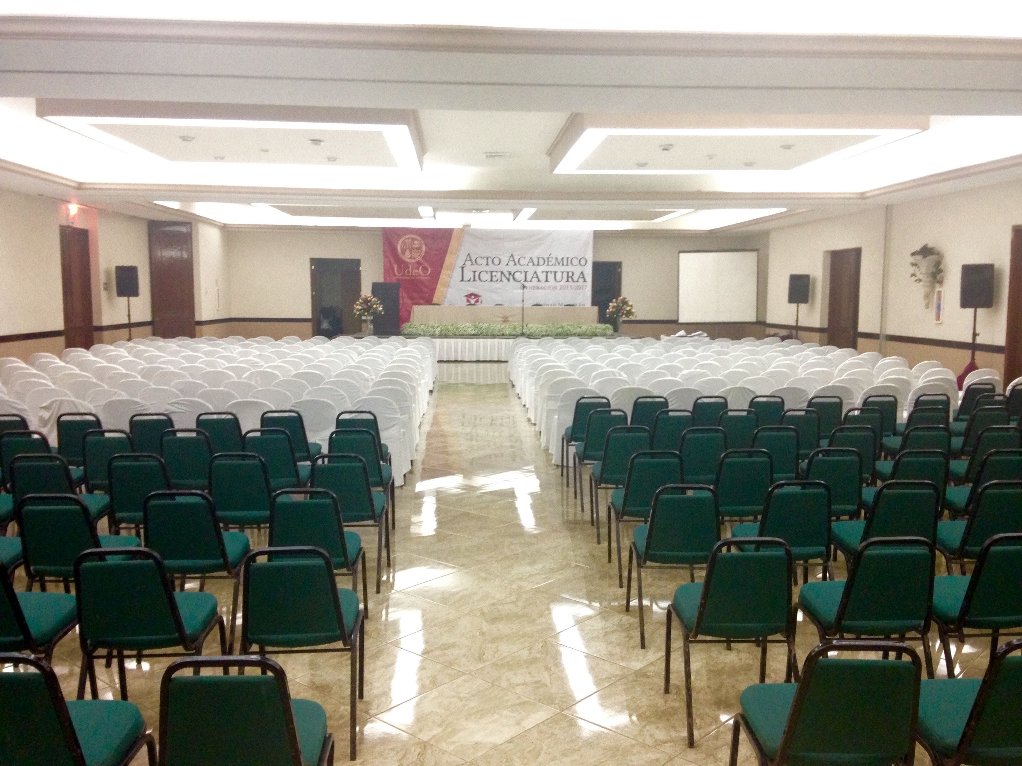 Un salón de conferencias con filas de sillas verdes y manteles blancos, un escenario con un banner y un suelo de baldosas.