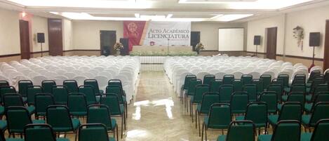 Un salón de conferencias con filas de sillas verdes y manteles blancos, un escenario con un banner y un suelo de baldosas.