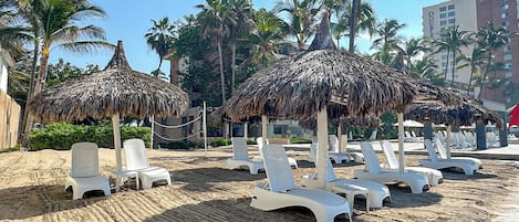 Una playa con sillas de playa blancas y sombrillas de paja, rodeada de palmeras y un edificio al fondo.