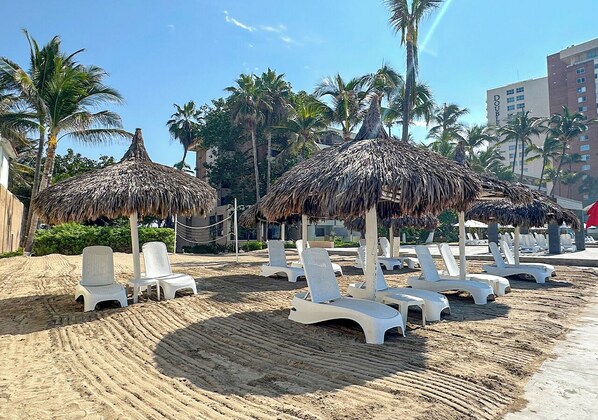 A beach with white lounge chairs and thatched umbrellas, surrounded by palm trees and a building in the background.