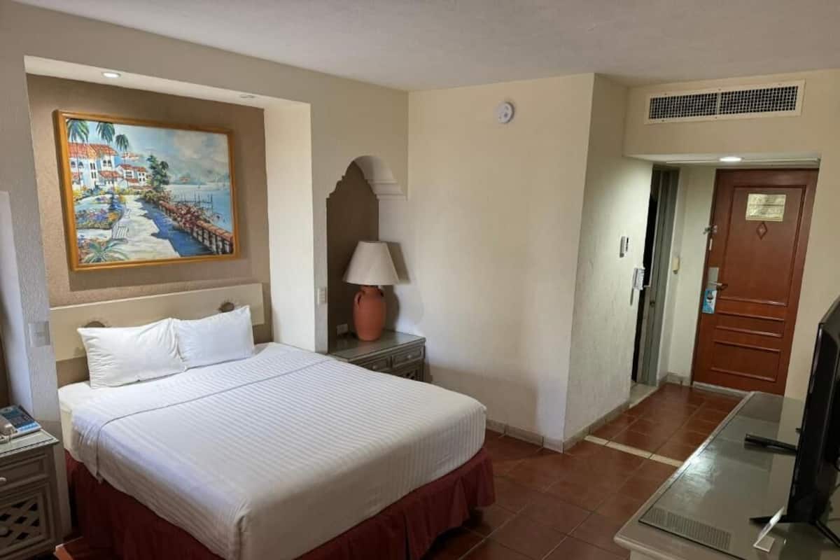 A hotel room with a bed, a nightstand, a lamp, a painting on the wall, and a door.
