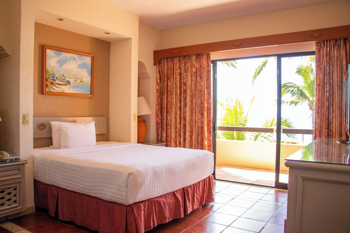 A hotel room with a bed, a painting on the wall, a lamp, and a view of the outdoors through a sliding door.