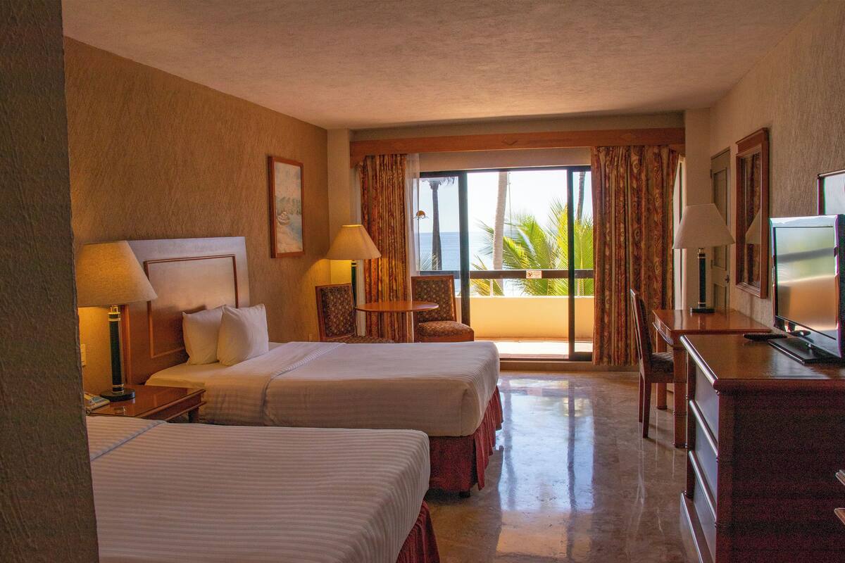 A hotel room with two beds, a television, a desk, and a balcony with a view of the ocean.