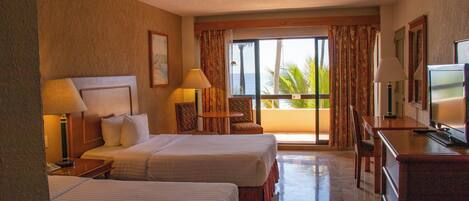 Habitación de hotel con dos camas, televisor, escritorio y balcón con vista al océano.