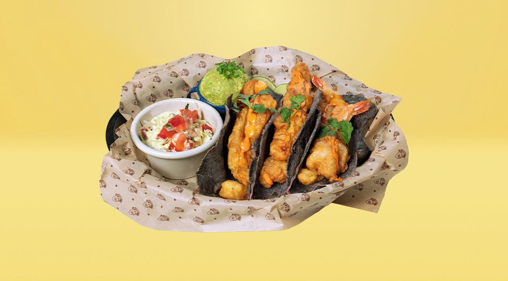A basket containing three fish tacos, a bowl of salsa, and a bowl of guacamole.