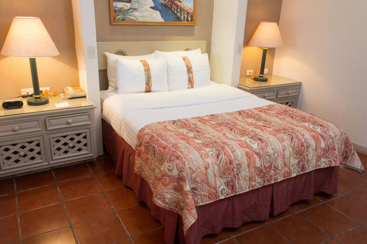A hotel room with a bed, two bedside tables, a nightstand, a lamp, a painting on the wall, and a tiled floor.