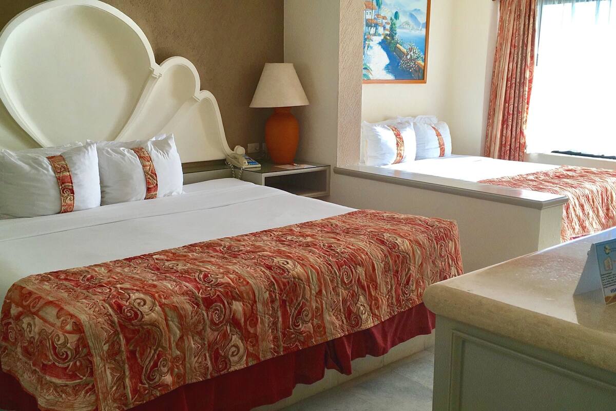 A hotel room with a large bed, a bedside table with a lamp, a painting on the wall, and a window with curtains.