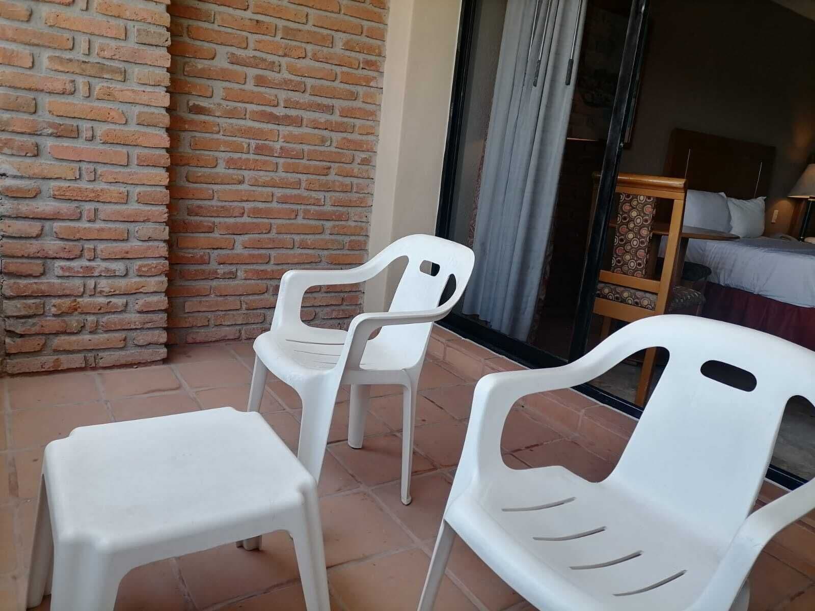 A patio area with a brick wall, two white plastic chairs, and a bed with a patterned headboard in the background.