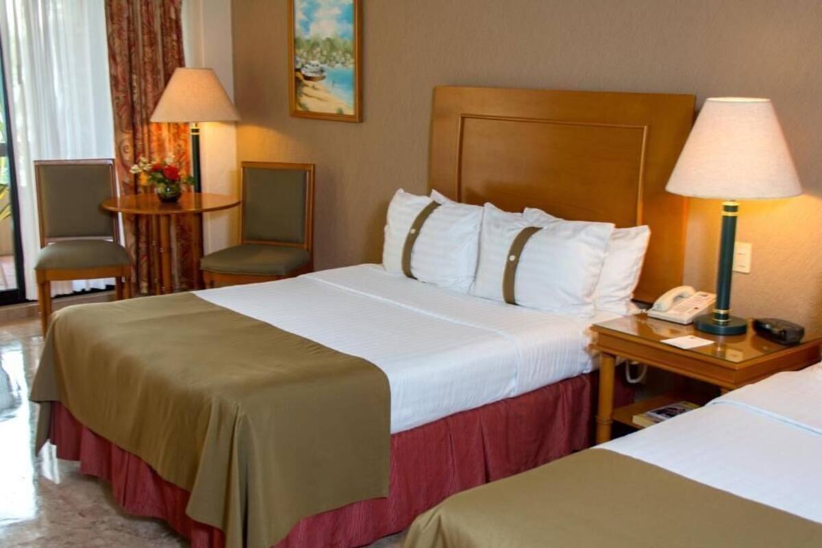 A hotel room with a bed, bedside tables, lamps, a chair, and a painting on the wall.