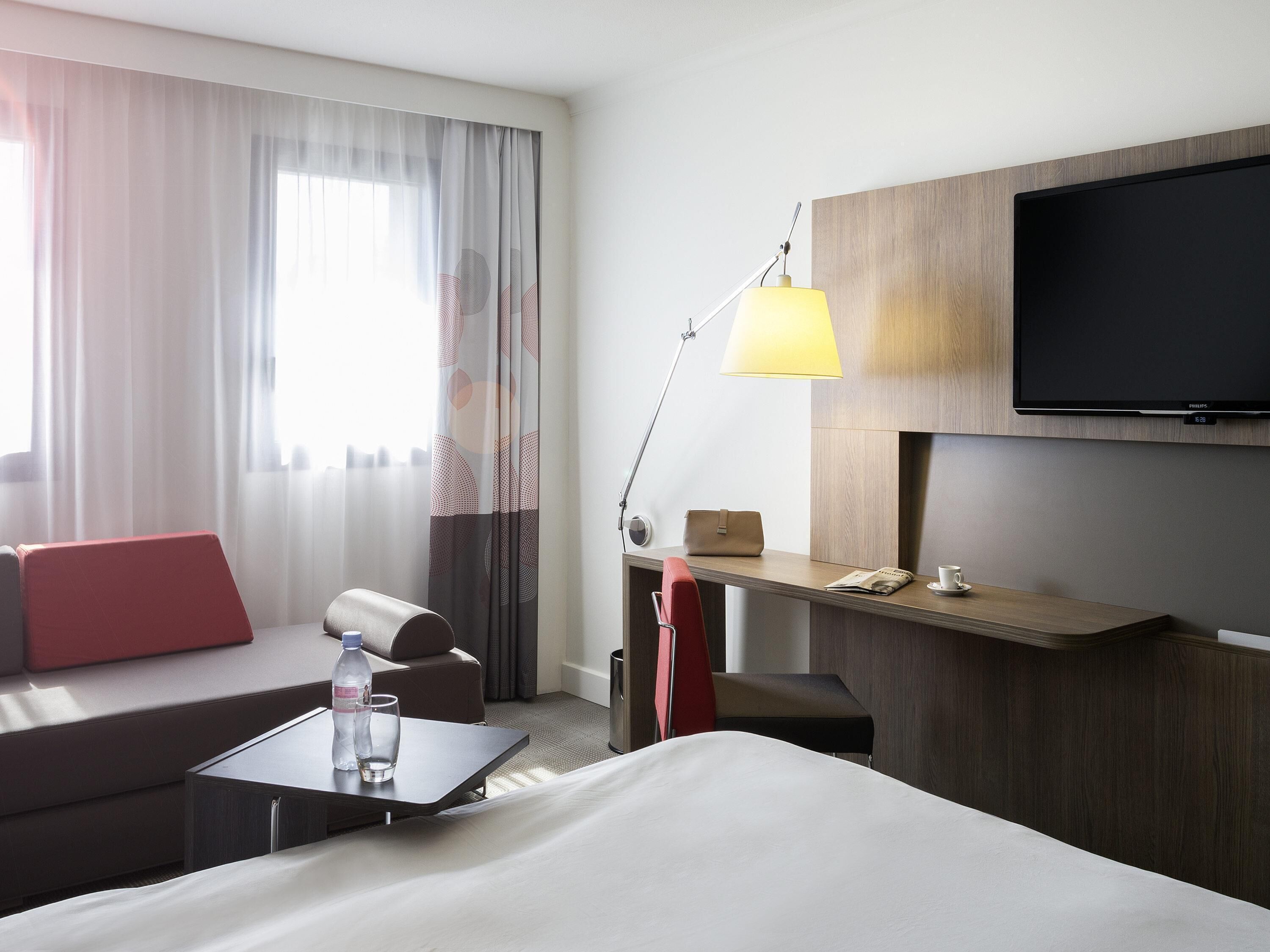 Ein Hotelzimmer mit Bett, Schreibtisch mit Lampe, Fernseher, Fenster mit Vorhängen und einem roten Sessel.