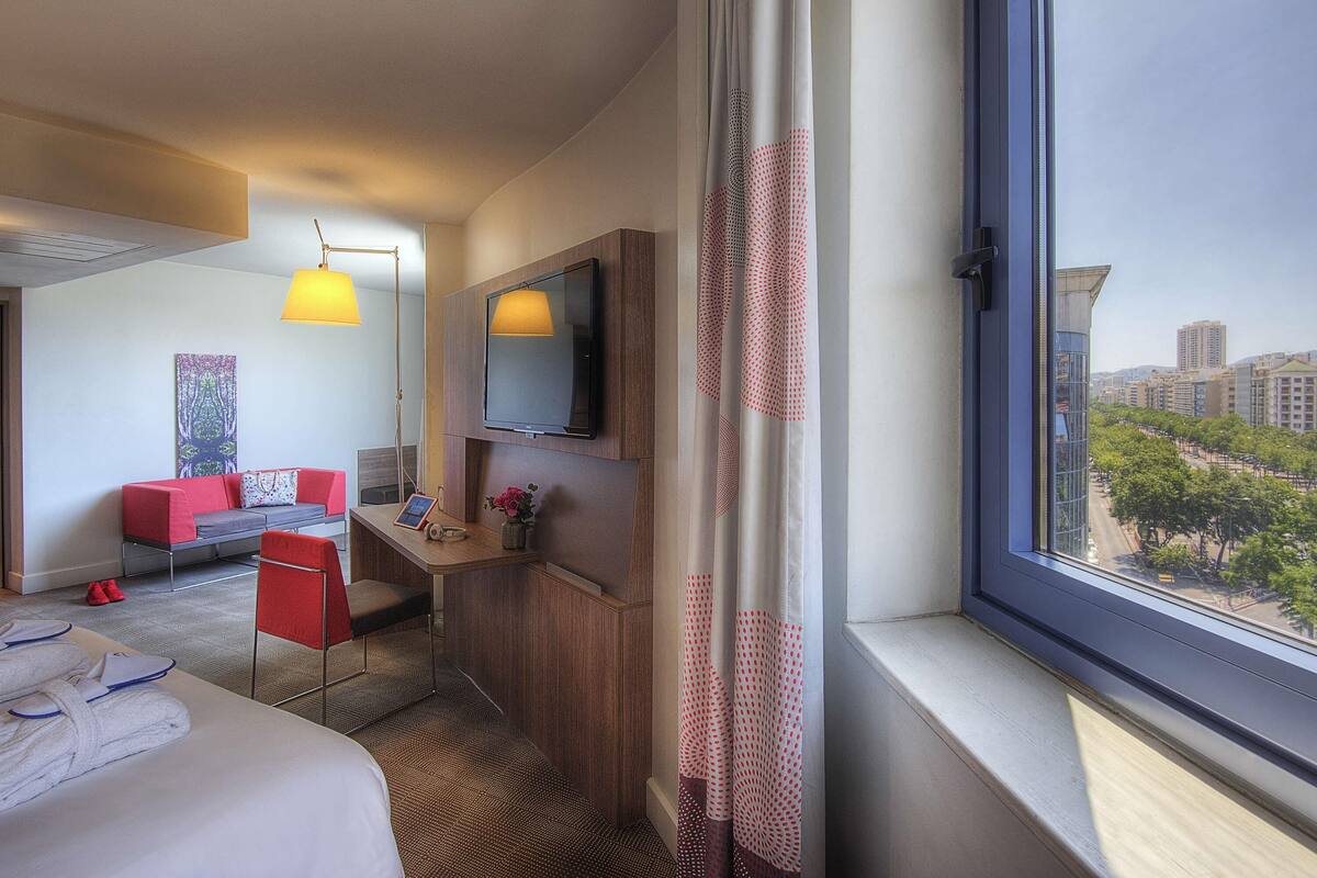 Een hotelkamer met een bed, een bureau met een computer, een televisie en uitzicht op de stad.