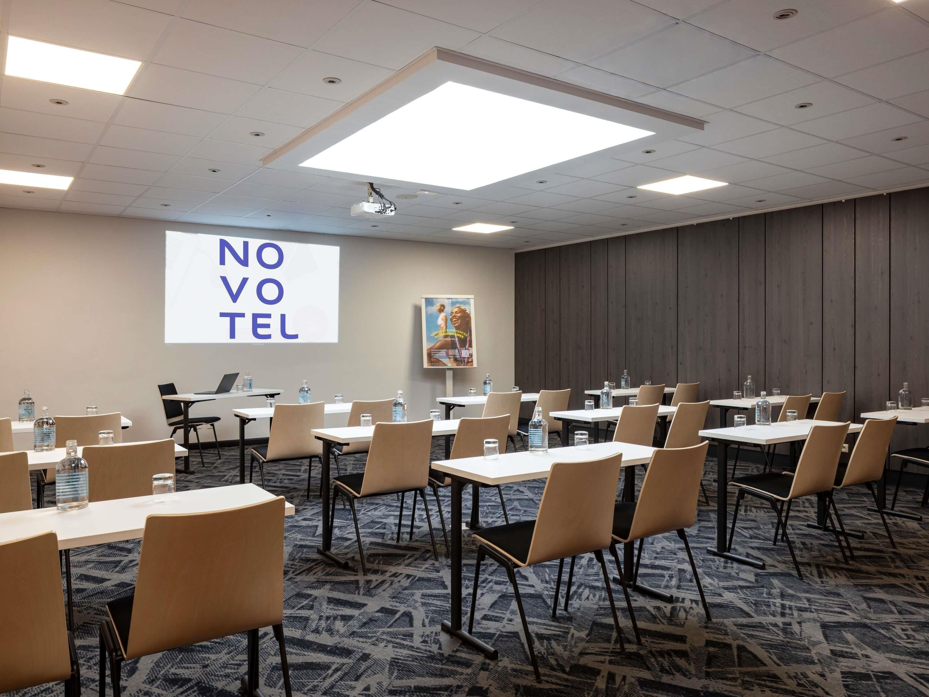 A conference room with tables, chairs, a projector screen, and a large 'NOVOTEL' sign.