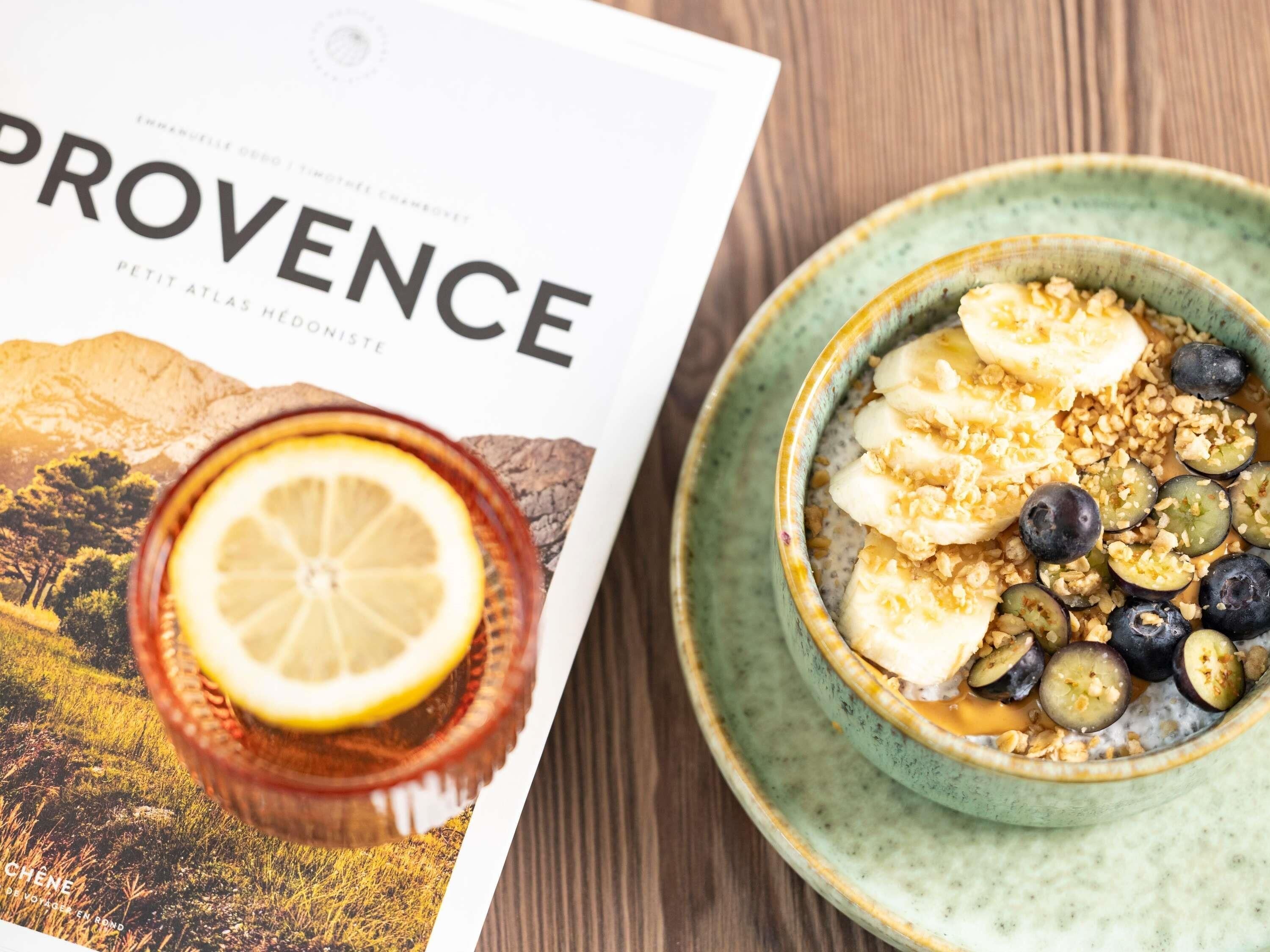A magazine with the word 'PROVENCE' on the cover, a bowl of oatmeal with bananas, blueberries, and granola, and a glass of orange juice with a lemon slice.