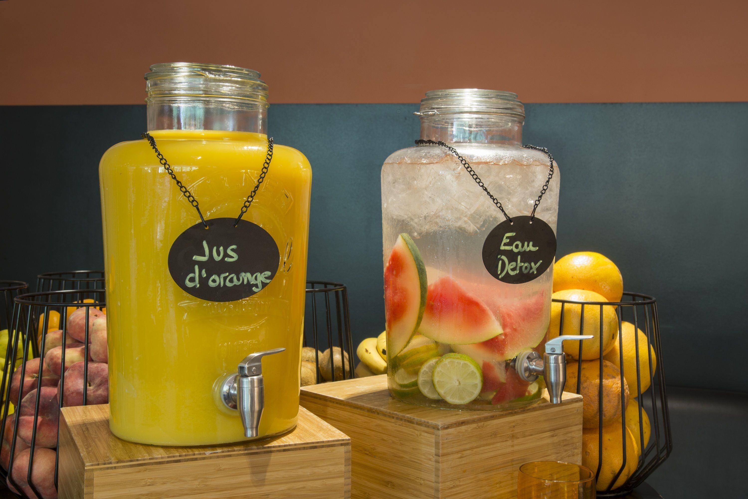 Two beverage dispensers, one labeled 'Jus d'orange' and the other 'Eau Detox', with fruits and ice visible inside.