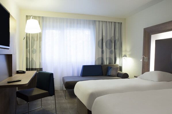 Una habitación de hotel con cama, escritorio, silla y un amplio ventanal con cortinas.