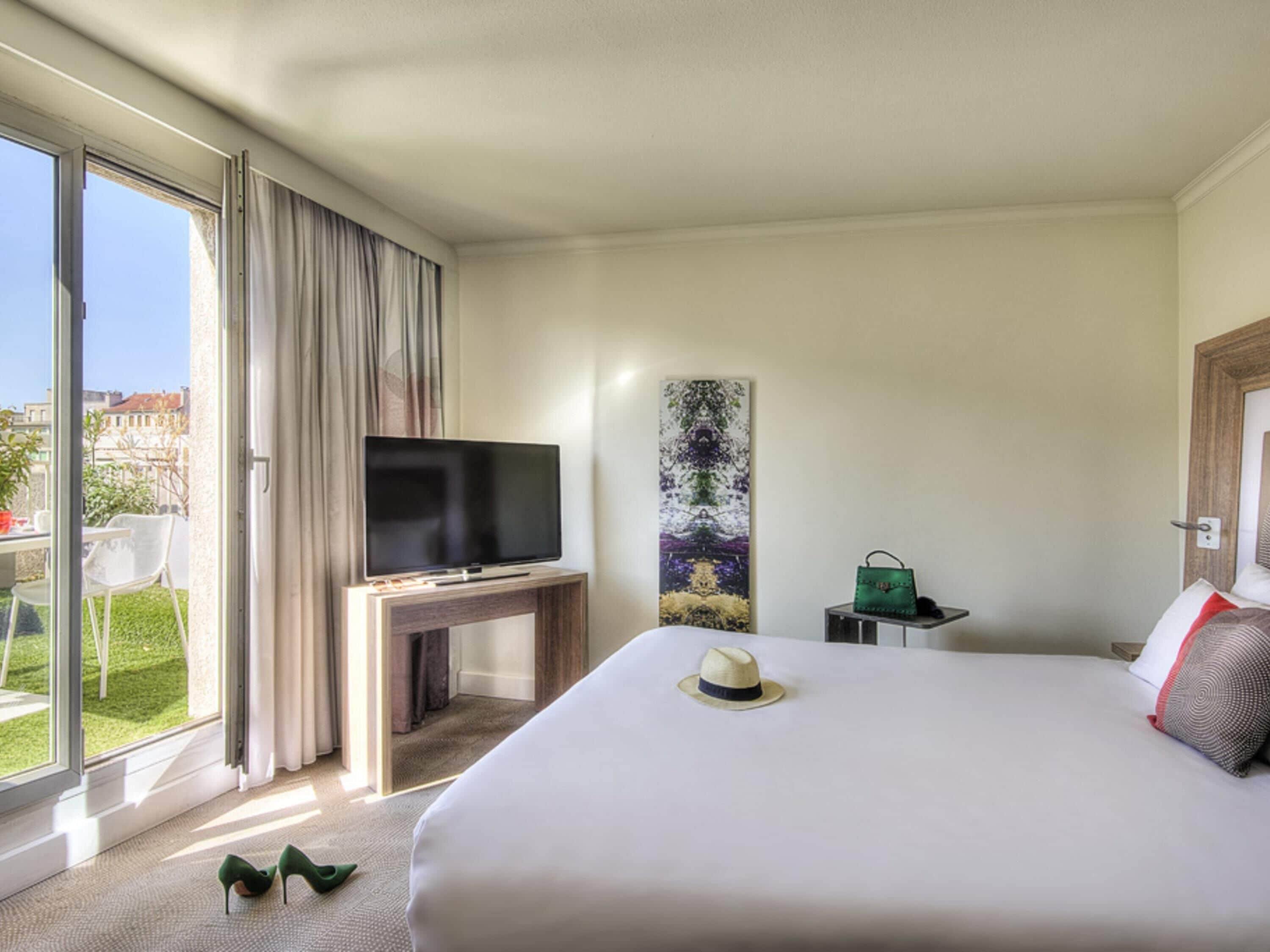 Une chambre d’hôtel avec un lit, un téléviseur posé sur un meuble, un tableau accroché au mur, une valise et une vue sur l’extérieur.