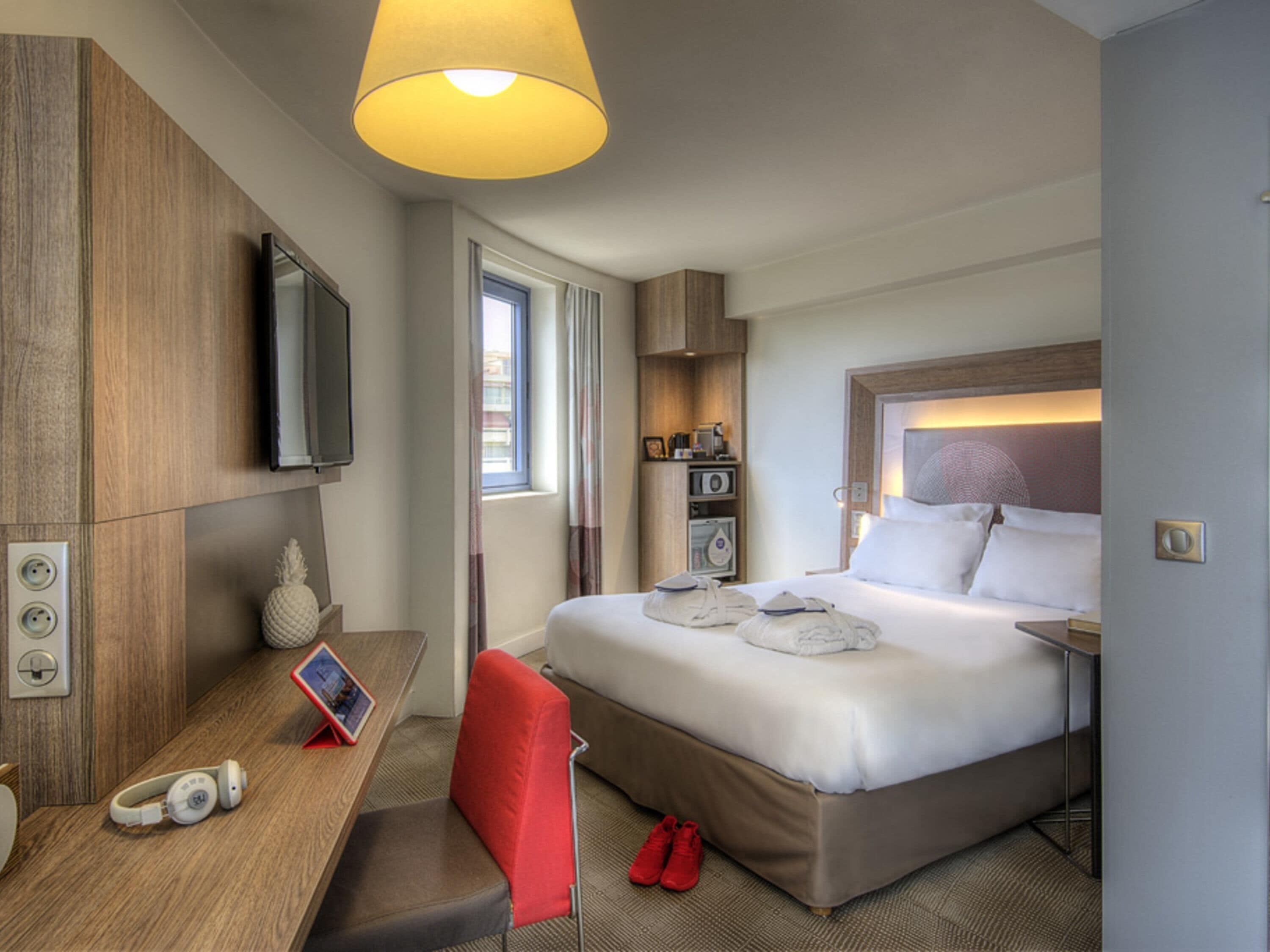 A hotel room with a bed, a desk with a chair, a television, a lamp, and a window with curtains.