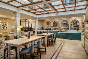 A spacious café with wooden tables and chairs, a bar area with a menu board, and a brick wall. - The Tremont House, Galveston, a Tribute Portfolio Hotel (Galveston)