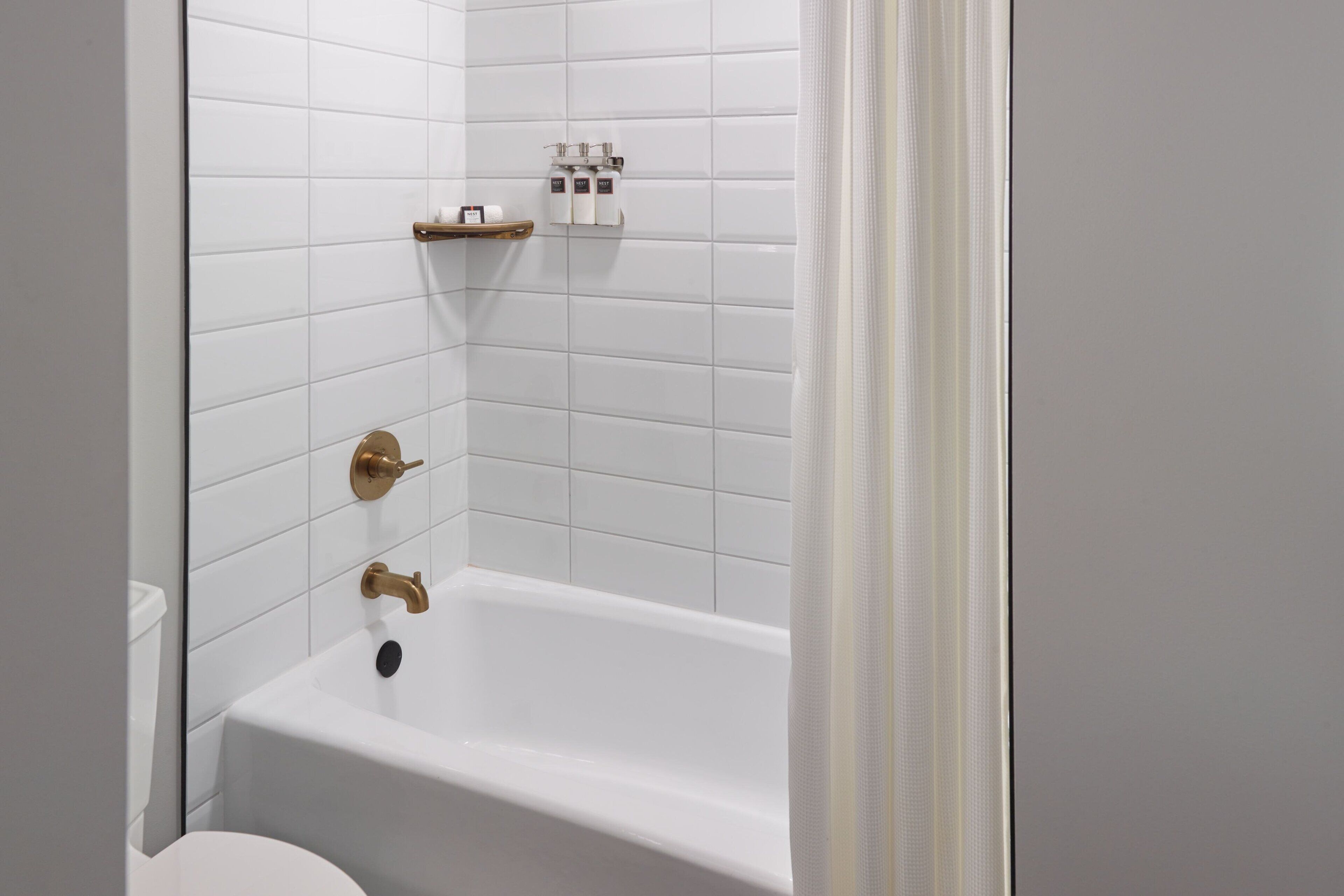 Un baño con paredes de azulejos blancos, una bañera blanca con detalles dorados y un estante con artículos de tocador.
