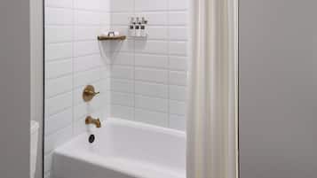 Un baño con paredes de azulejos blancos, una bañera blanca con detalles dorados y un estante con artículos de tocador.