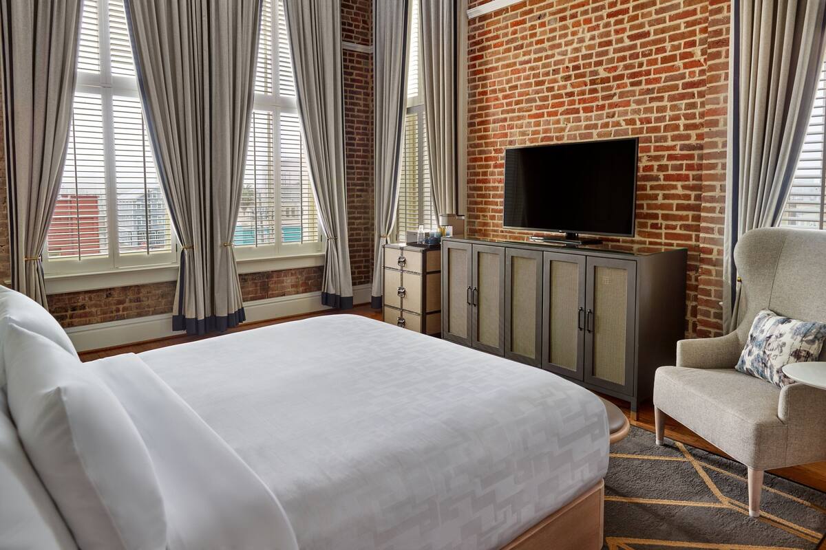 A hotel room with a large bed, a TV on a brick wall, a grey armchair, and a window with curtains.