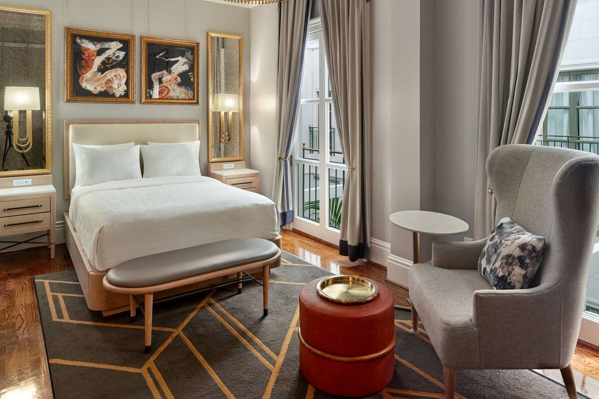 A hotel room with a large bed, a bedside table with a lamp, a chair with a pillow, a small round table, and a window with curtains.