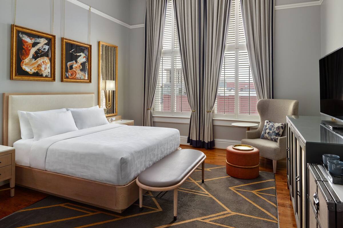 A hotel room with a bed, a bench, a TV, a desk, and two paintings on the wall.