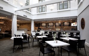 A spacious indoor dining area with multiple tables and chairs, a brick wall with menu boards, and a television screen mounted on the wall. - The Tremont House, Galveston, a Tribute Portfolio Hotel (Galveston)