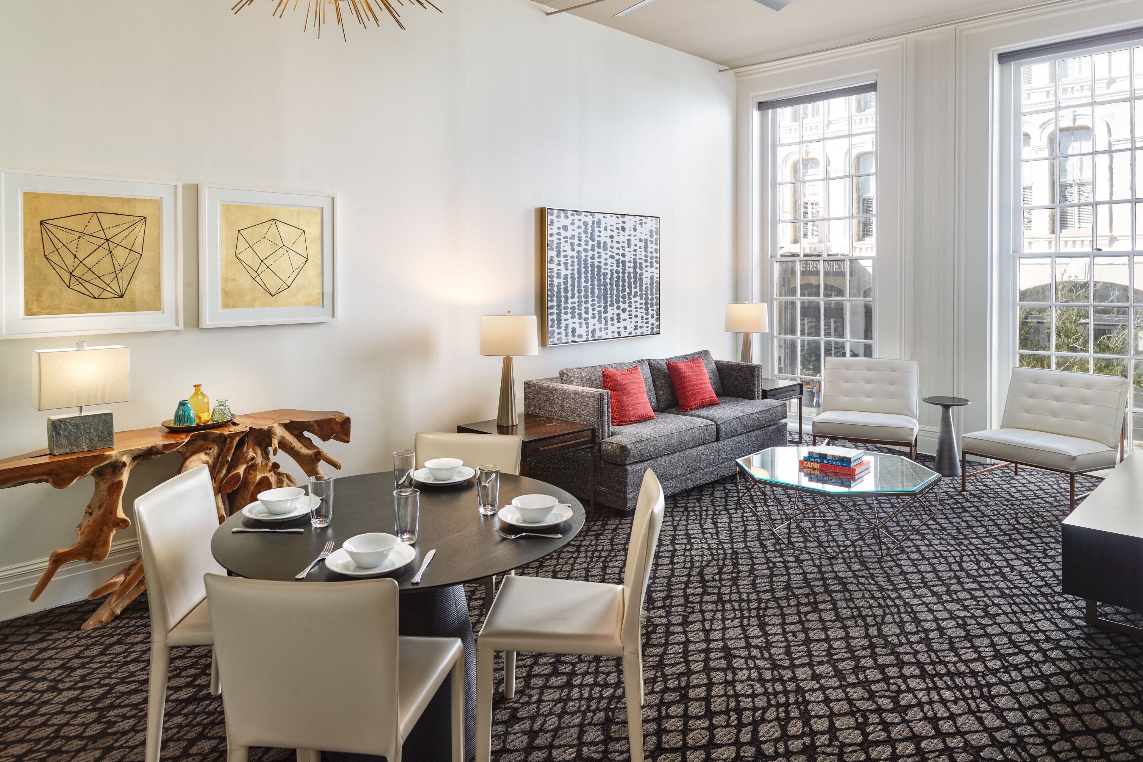 Une chambre d’hôtel moderne avec un coin repas, un canapé et une table basse. Les murs sont ornés d’œuvres d’art encadrées, il y a une grande fenêtre et un tapis à motifs.