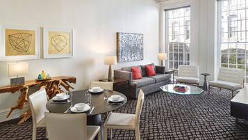 Una habitación de hotel moderna con zona de comedor, sofá y mesa de centro. Se observan obras de arte enmarcadas en la pared, un ventanal grande y una alfombra estampada.