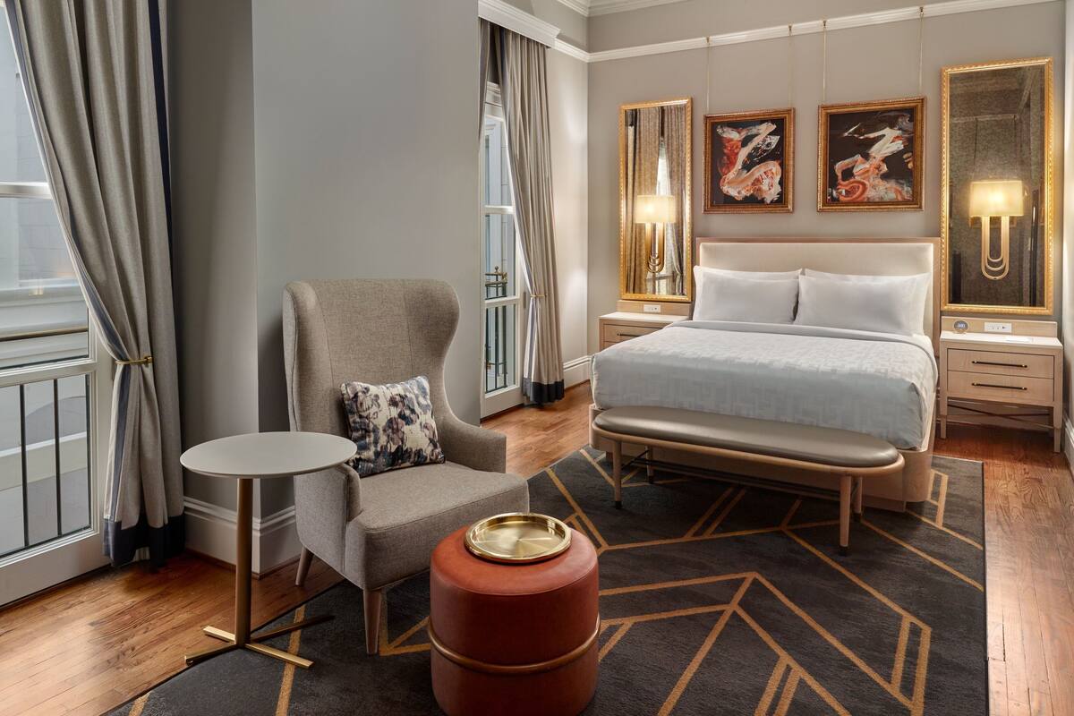 A hotel room with a bed, a chair, a small table, and a large mirror.