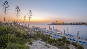 A coastal view with a sunset, beach chairs, umbrellas, and a distant building. - Corinthia St George's Bay (St. Julian's)