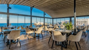 A spacious dining area with tables and chairs, large windows offering a view of the sea and cityscape. - Corinthia St George's Bay (St. Julian's)
