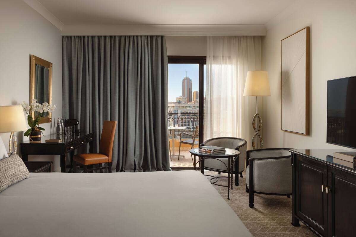 Ein Hotelzimmer mit Bett, Schreibtisch mit Lampe, Sessel, Fernseher und Blick auf die Stadt.