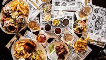 Una mesa con diversos platos, incluyendo pescado con papas fritas, patatas fritas y alitas de carne, acompañados de salsas y bebidas.