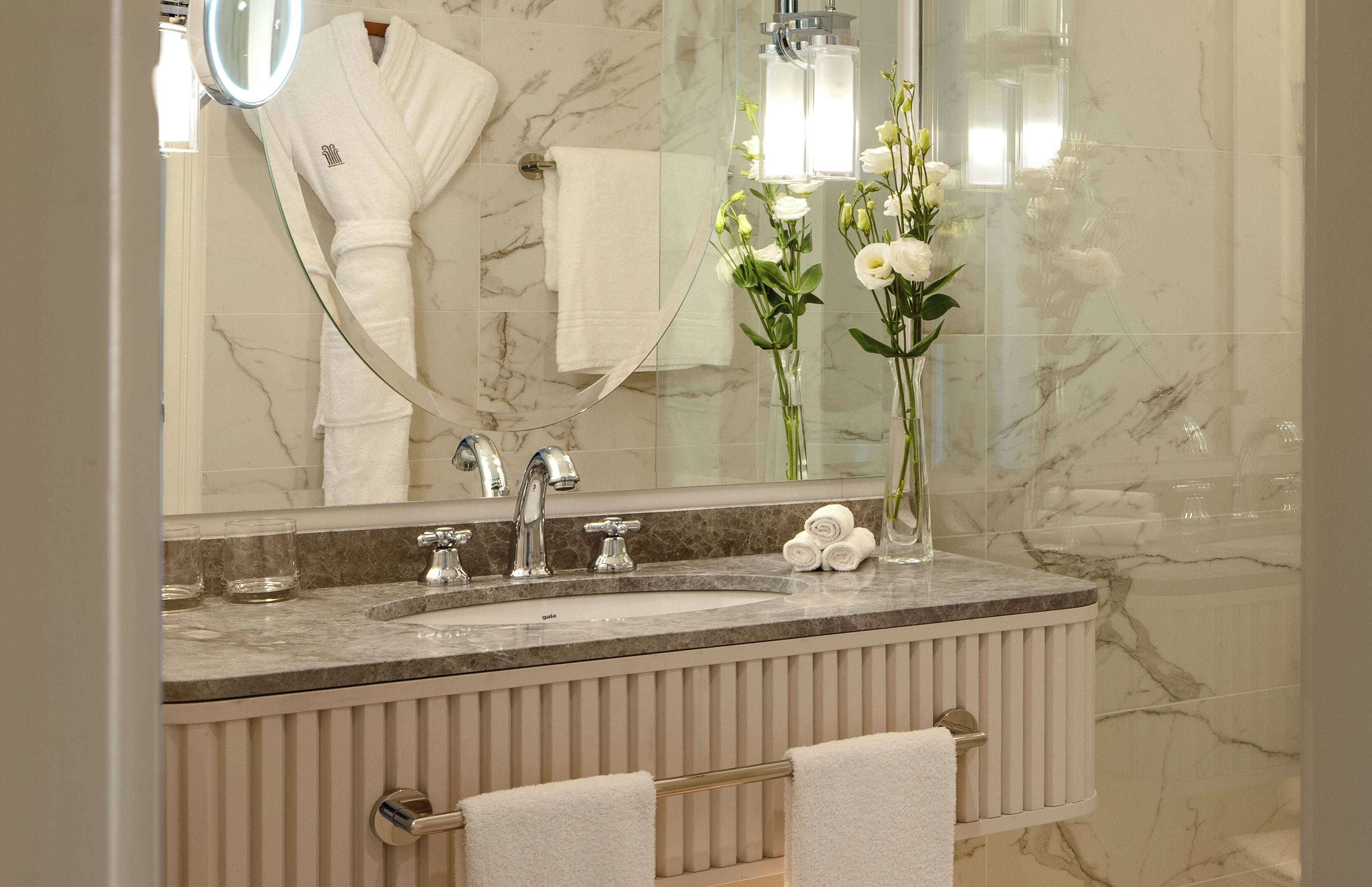 A bathroom with a marble sink, two faucets, a mirror, a towel rack with towels, and a vase with flowers.