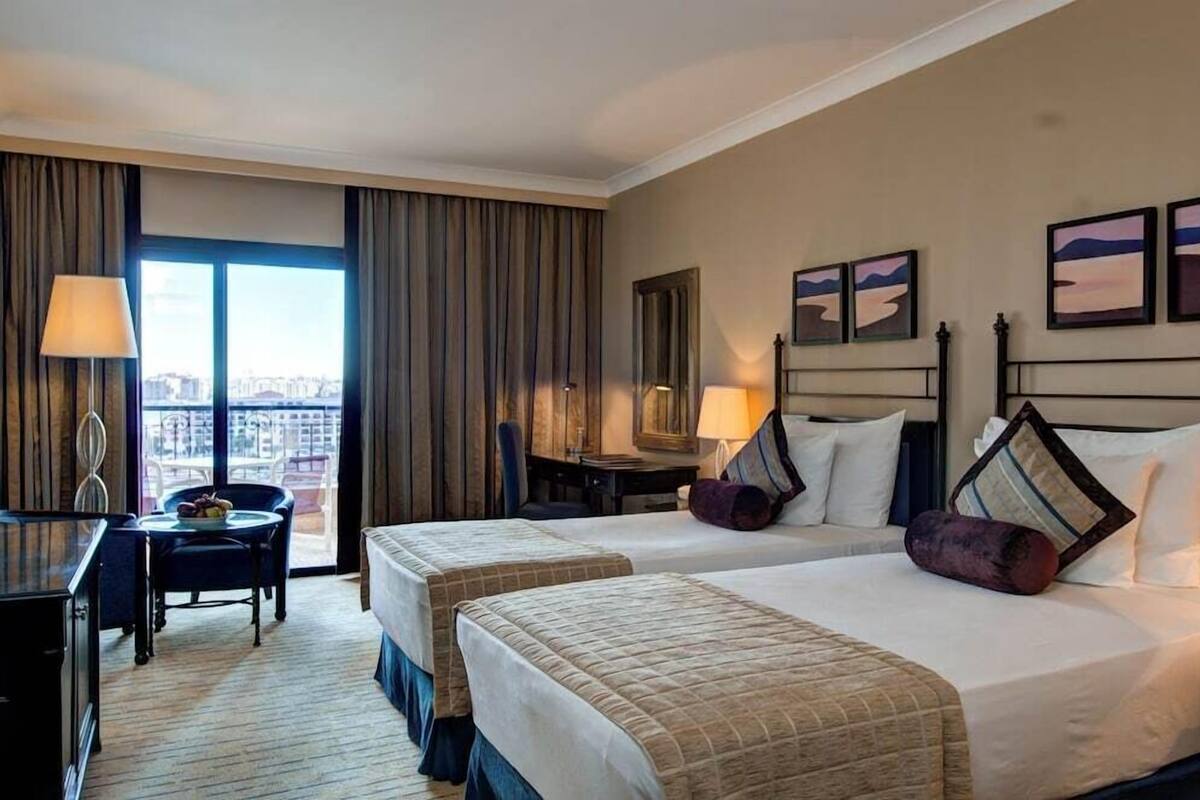 Ein Hotelzimmer mit zwei Betten, einem Schreibtisch, einem Sessel und einer Lampe, mit Blick auf die Stadt durch große Fenster.