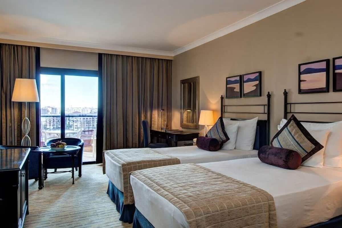 Une chambre d’hôtel avec deux lits, un bureau, une chaise, une lampe et une vue sur la ville grâce à de grandes fenêtres.