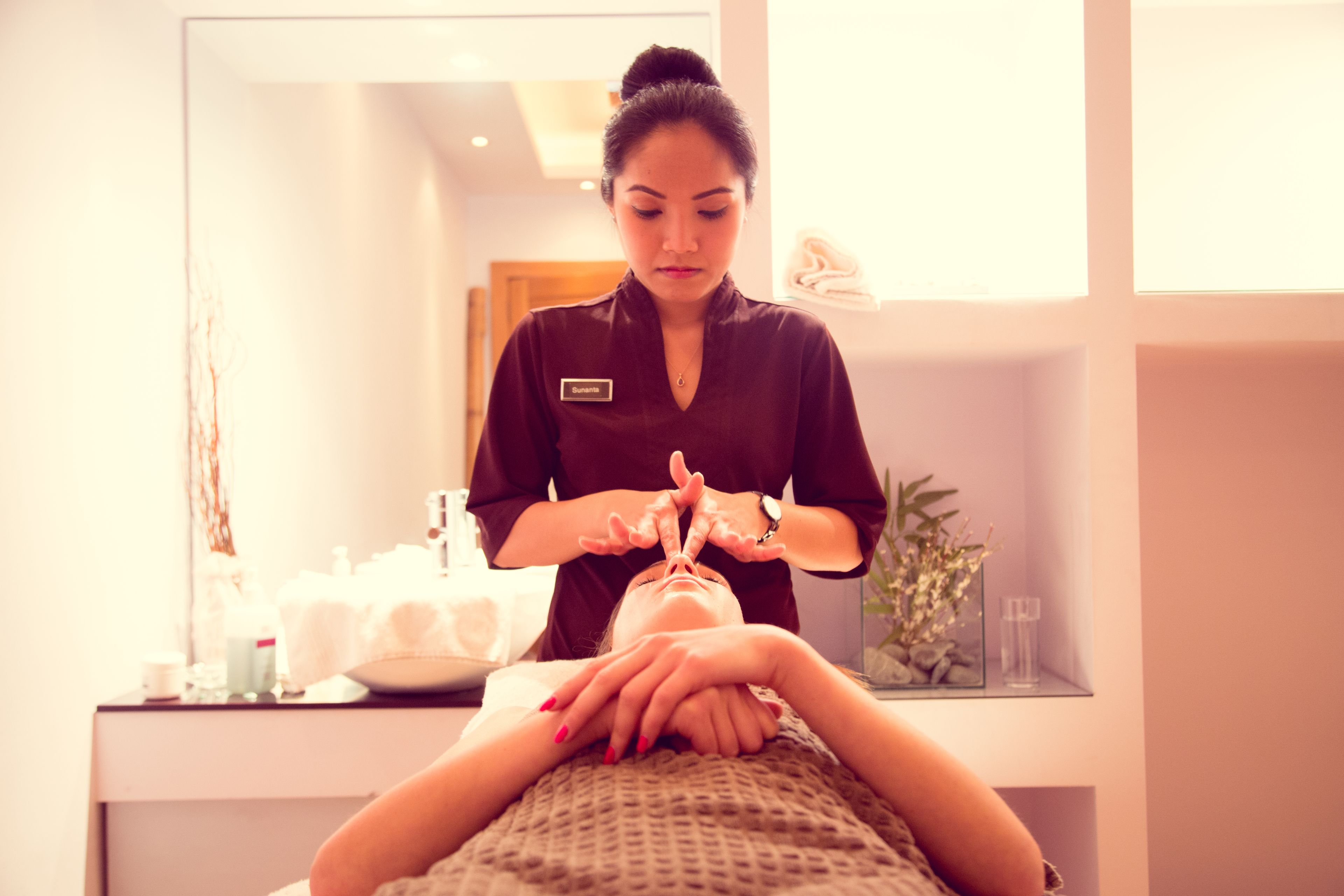 A person receiving a facial treatment in a well-lit room with a mirror, a sink, and a plant.