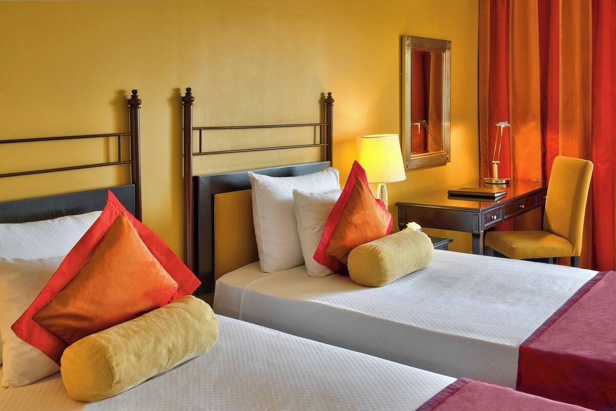 Ein Hotelzimmer mit zwei Betten, jeweils mit weißer Bettwäsche und roten und beigen Kissen. Es gibt einen Schreibtisch mit einer Lampe, einen Stuhl und einen Spiegel an der Wand.