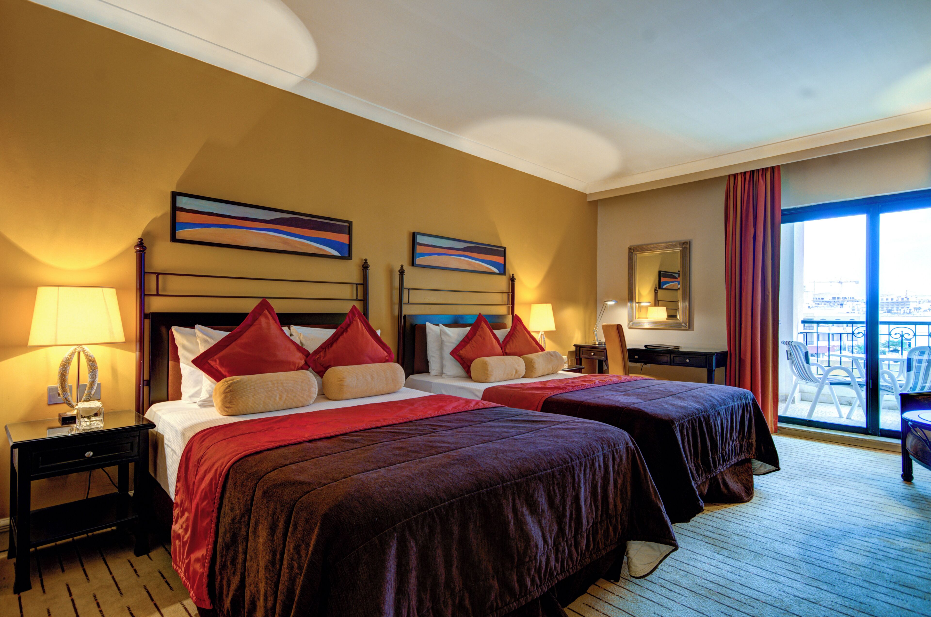 A hotel room with two beds, a desk, a chair, and a balcony with a view.