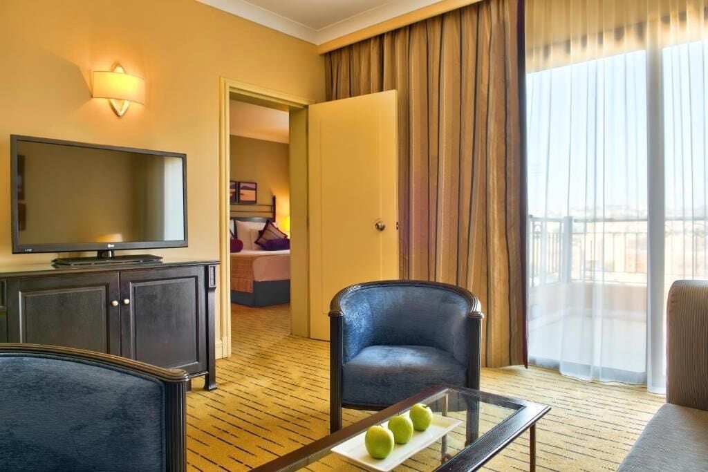 Quarto de hotel com televisão de tela plana, poltrona escura, mesa de centro de vidro com maçãs, piso de madeira e uma janela grande com cortinas.