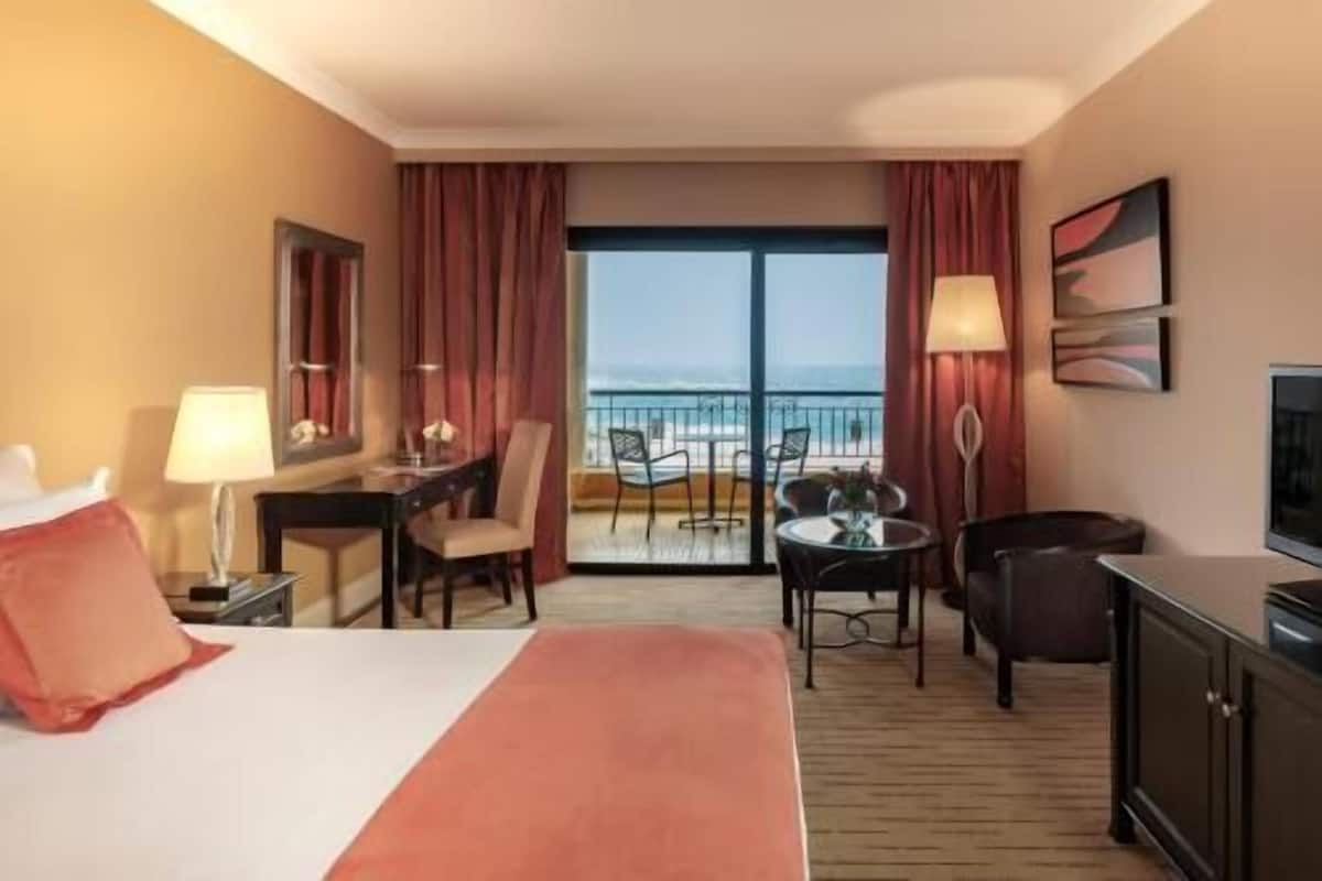 Ein Hotelzimmer mit Bett, Schreibtisch, Stuhl und Blick auf den Strand.