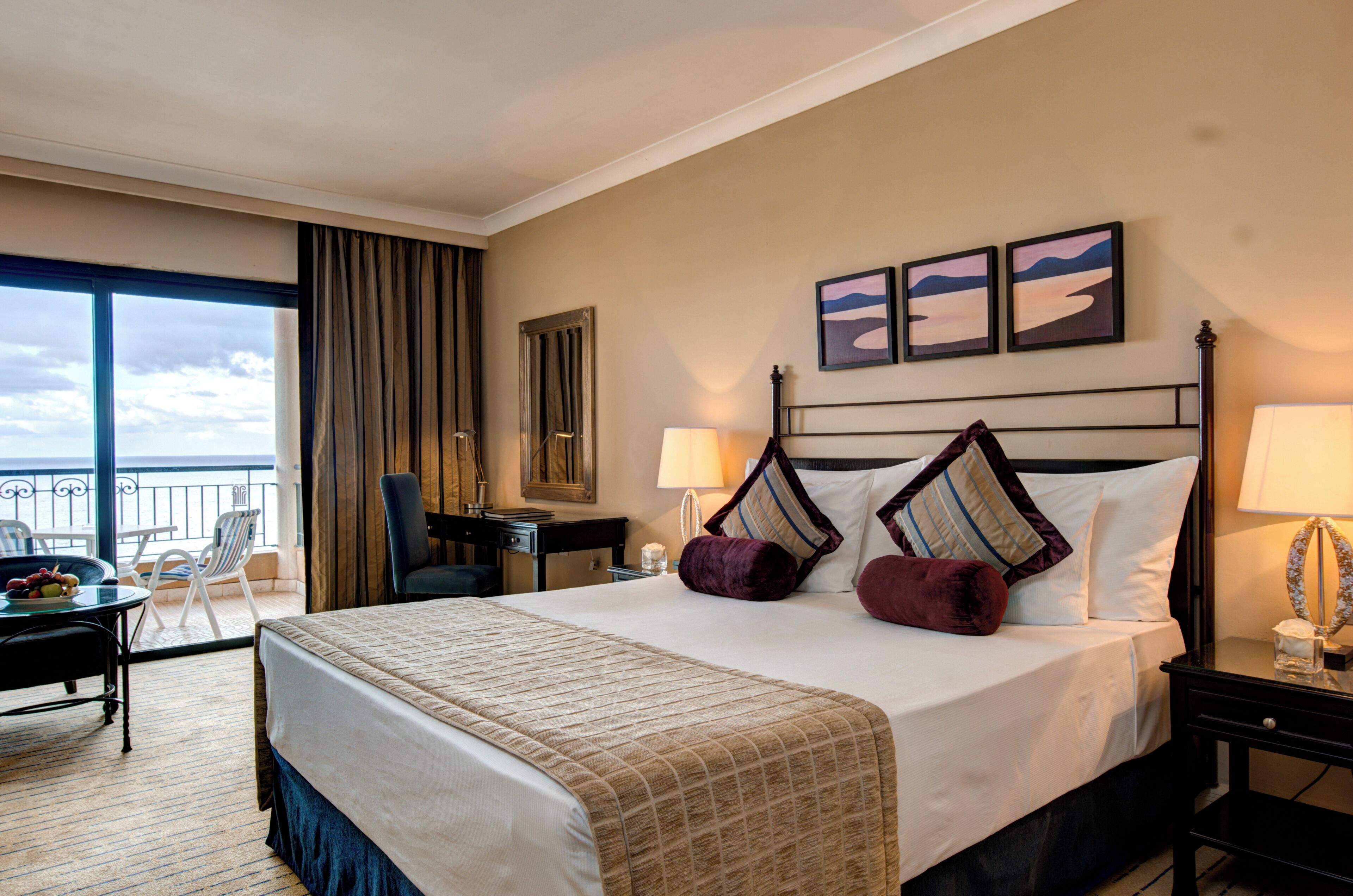 A hotel room with a large bed, two bedside tables, a desk with a chair, a balcony with a view, and artwork on the wall.
