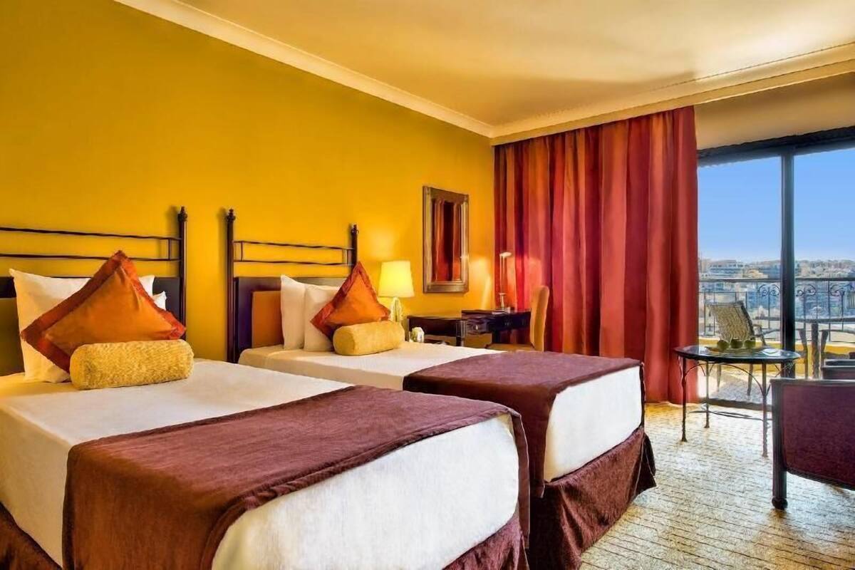 Ein Hotelzimmer mit zwei Betten, einem Schreibtisch, einem Stuhl und einem großen Fenster mit Blick auf die Stadt.